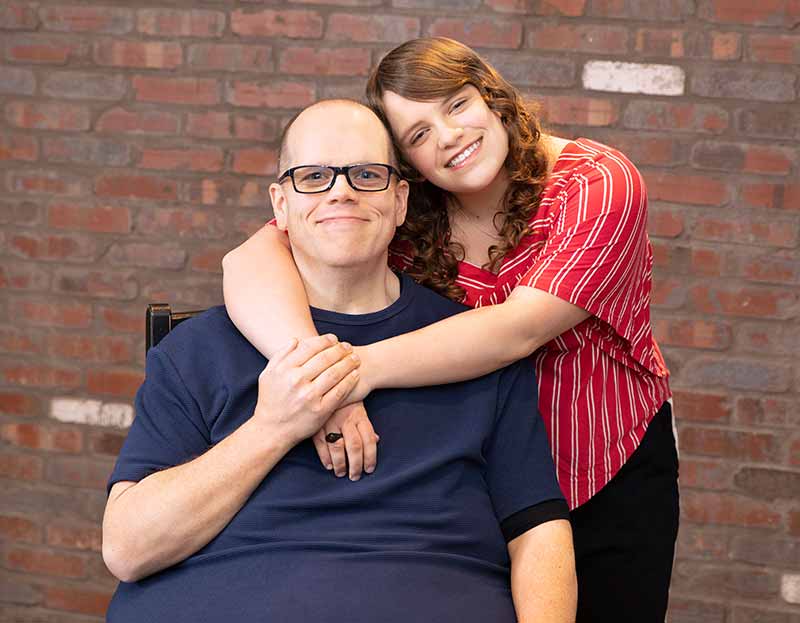 Dennis Guzy, who wears a T-shirt and has glasses and a crewcut, sits in a chair in front of a brick wall. His daughter, Mikala, who has long, curly hair and wears a striped shirt, stands beside him with her arms around his shoulders.