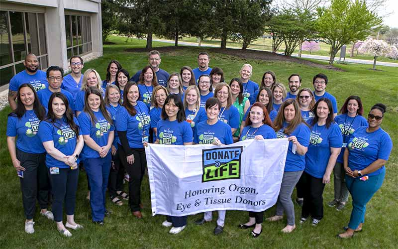 A group photo on a grassy lawn with a building and trees in the background. Everyone is in matching t-shirt with the 'Donate Life' icon on the front, paired with either denim or black pants. In front of the group, four people hold a white banner with the 'Donate Life' emblem and the words 'Honoring Organ, Eye, and Tissue Donors'.