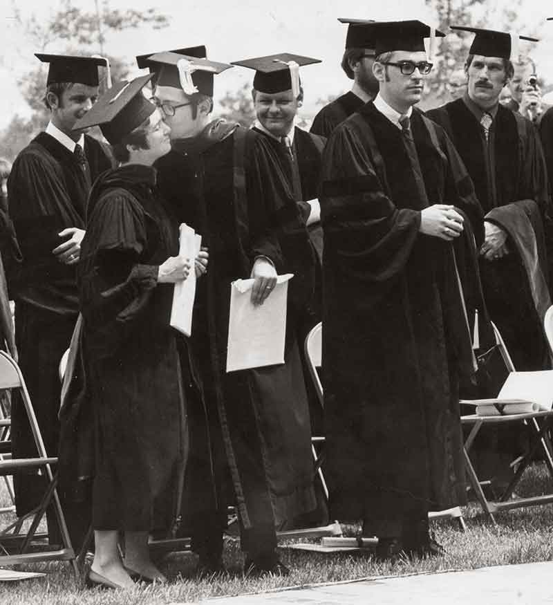 Eight medical students stand and wear caps and gowns at Penn State College of Medicine’s first graduation ceremony in 1971. Folding chairs are behind each of them. On the left, a man kisses a woman on the cheek.  Penn State College of Medicine’s first class graduates in 1971.