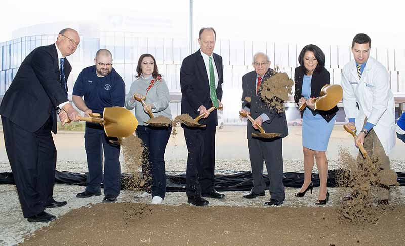 Seven people stand in a row using ceremonial shovels to toss dirt in the air at the Penn State Health Hampden Medical Center groundbreaking. Behind them is a large construction plan rendering of the new hospital on March 29, 2019. From left are Dr. Craig Hillemeier, former dean of Penn State College of Medicine, chief executive officer of Penn State Health and Penn State’s senior vice president for health affairs, an unidentified man and woman from the community; Alan Brechbill, executive vice president and chief operating officer for Penn State Health; Al Beinstock, a former Hampden Township commissioner; Deb Rice-Johnson, president of Highmark Inc.; and Dr. James Tucker with Penn State Health Medical Group ― Camp Hill.