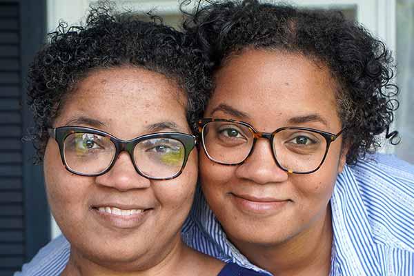 Twin sisters Alana and Alicia Webb smile as they pose in front of a window at their home. Both women have short curly hair and wear glasses.