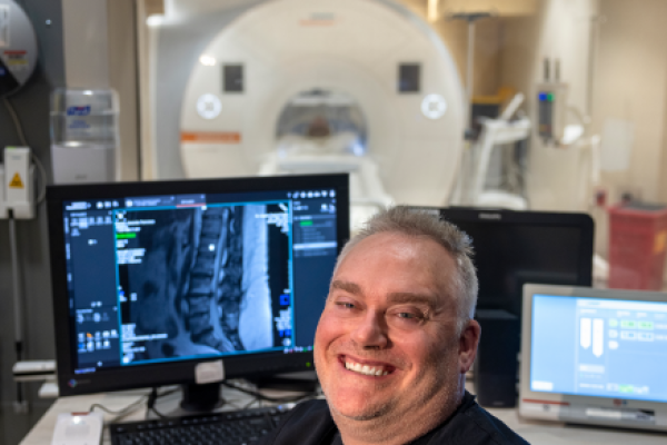 Gajewski is seated at his desk facing the camera with an MRI scan visible on his computer screen. Behind him in the lab is the MRI machine.