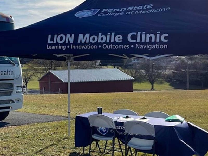 Penn State College of Medicine LION Mobile Clinic tent and table is in an open grassy field. The Mobile Health van is pictured to the left of the tent. of the 