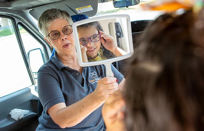 Nancy Mesko, optician with Vision to Learn, holds a mirror in front of Gabriella, whose image in seen in the mirror as she checks how a pair of glasses look on her face. Mesko has short hair and glasses and wears a T-shirt with a Vision to Learn logo on it.