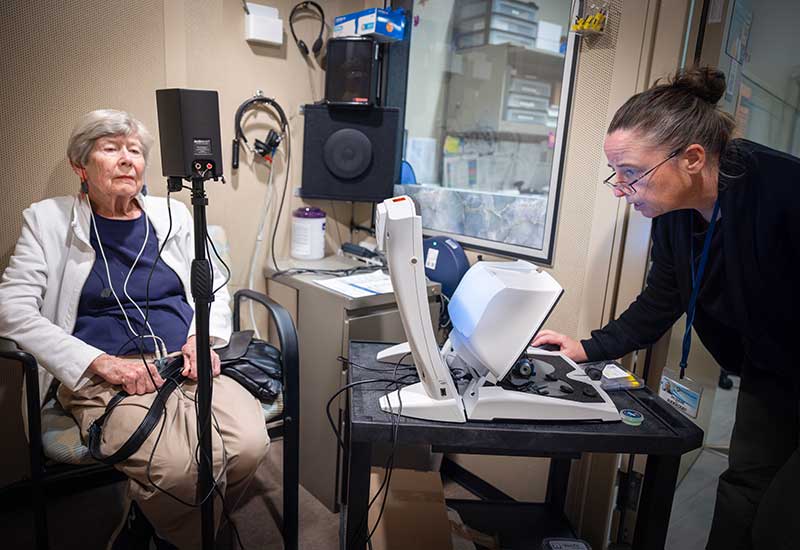 Person undergoing a hearing test in a soundproof room while another operates audiology equipment on a desk.