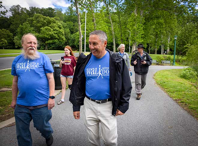 Two men wearing Walk With a Doc T-shirts smile as they walk at City Park in Reading. Behind them, three other people walk along a path.
