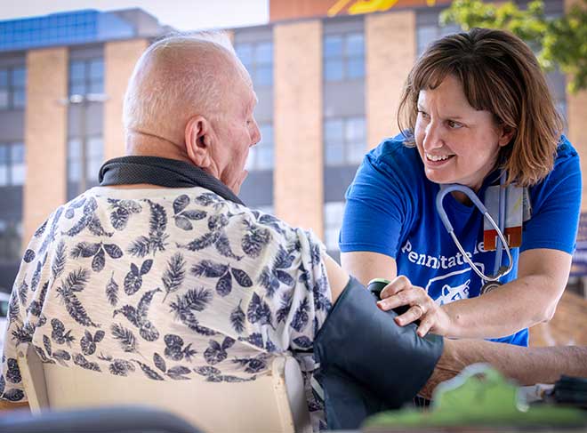 A community health nurse at Penn State Health smiles at a man as she checks his blood pressure. She is wearing a T-shirt and has a stethoscope around her neck. He is wearing a shirt with a leaf print on it.