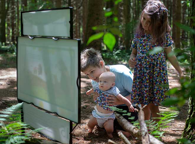 A boy holds a baby up in front of a sign. On their right is a three-year-old girl wearing a dress. They are on a nature trail.