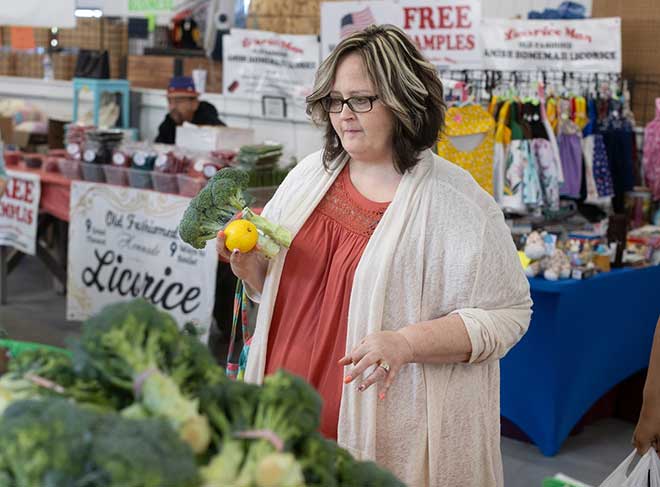 A woman holds a head of broccoli while looking at other vegetables at a farmer’s market. She is wearing glasses and a sweater.
