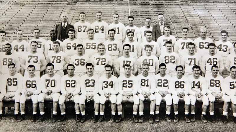 The 1948 Penn State football team poses for a photo. Four rows of players sit or stand. The coach wearing a suit and tie stands in the top-left row.