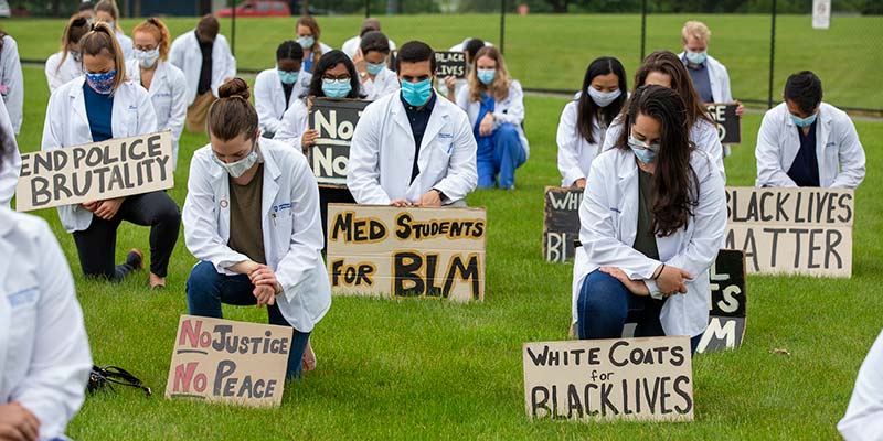 Photo of a group of people wearing white medical coats and masks, kneeling on the grass and holding signs that say “Black Lives Matter,” “White Coats for Black Lives,” “End Police Brutality,” and “No Justice, No Peace.”