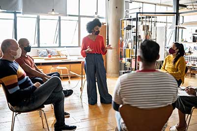 Employees wearing masks sit in circle listening to one employee who is standing and speaking.