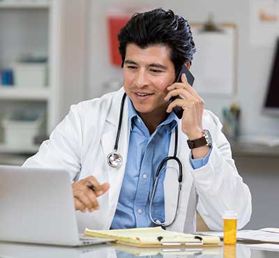 Doctor wearing lab coat and stethoscope sits at desk and reviews computer screen while talking on cell phone.