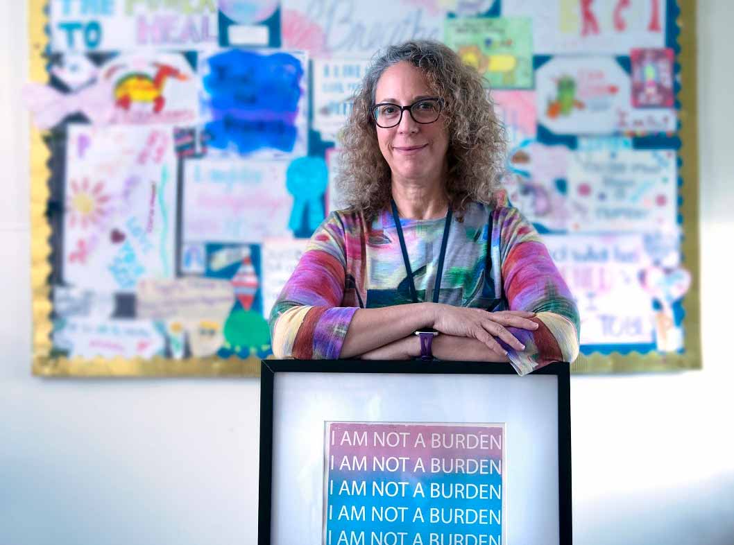 Amy Keisling, Gender Health Clinic coordinator, smiles as she leans on a framed picture that says “I am not a burden.” She has curly hair and is wearing glasses and a lanyard. Behind her is a bulletin board out of focus.