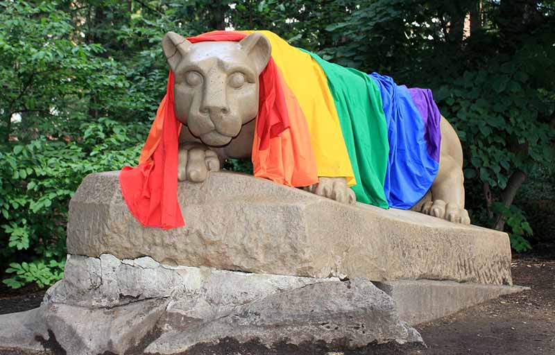 Photo of the Penn State Nittany Lion statue in front of some trees with an LGBTQ+ Rainbow Pride flag draped over its back.