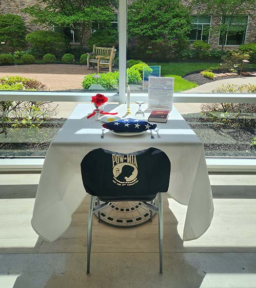 Photo of the Fallen Comrade Table includes a white table cloth, a single red rose with a red ribbon tied around the vase, a slice of lemon on a dish, a salt shaker, the Bible, an inverted glass. There is an empty chair at the table with a POW-MIA blanket placed over the chair back.