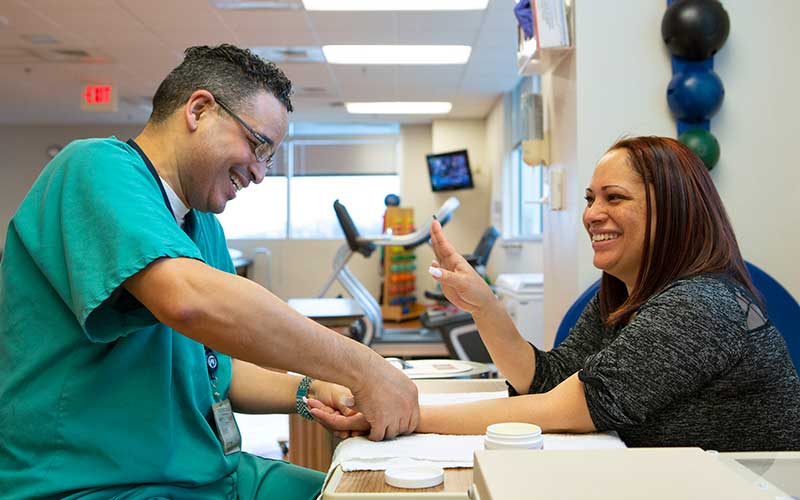 Julio Torres, an occupational therapist with Penn State Health St. Joseph Medical Center, works with a patient. He is seated at a workstation. A patient is seated on the other side of the workstation. A combination of therapy equipment is pictured in the background.