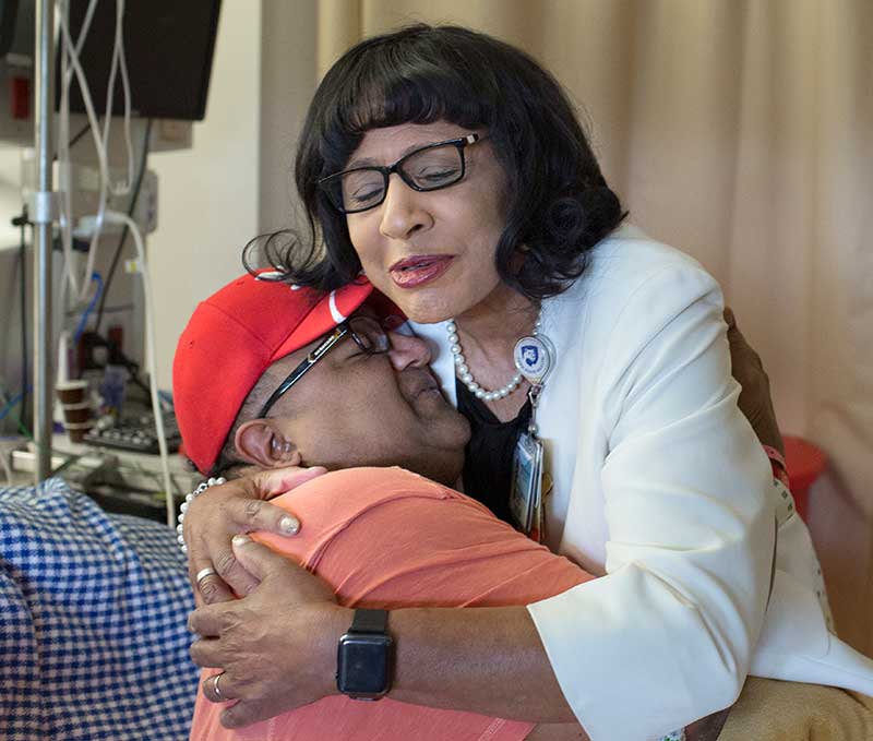 A chaplain gently embraces a person in a hospital room, with medical equipment and curtains visible in the background.