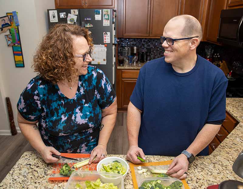 Michelle Guzy, who has short, curly hair and wears glasses and a patterned shirt, smiles at her husband, Dennis Guzy, as they stand at their kitchen counter cutting vegetables on cutting boards. Dennis wears a T-shirt and has glasses and a crew cut.