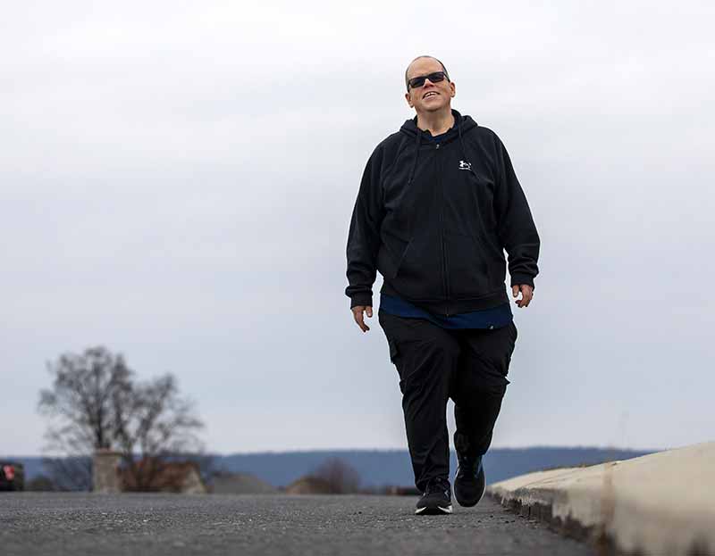 Dennis Guzy, dressed in a jacket, jogging pants and wearing sunglasses, walks along a gravel road. Parked and cars and trees are in the background