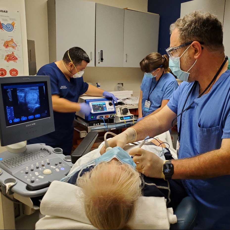 "Ear, Nose and Throat surgeon in blue scrubs holding two instruments up to the neck of a female patient. He is wearing glasses and a blue mask. There is a screen on the left that he is looking at as he is performing radiofrequency ablation on the patient’s thyroid nodules. There are two other staff members in the background, also in scrubs, monitoring equipment. "