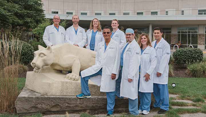 Eight surgeons gather around a statue of the Nittany lion that sits in a garden in front of Penn State Health St. Joseph Medical Center. They all are dressed in scrubs and doctor coats. Shrubbery is visible in the garden around them. A small tree is in the back left of the garden.
