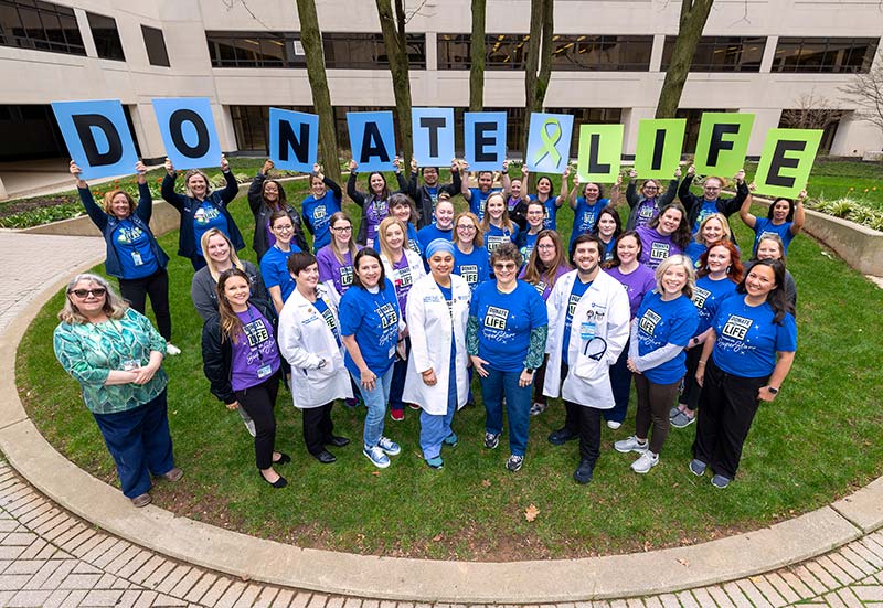 A group photo on a grassy lawn with a building and trees in the background. Everyone is in matching t-shirt with the 'Donate Life' icon on the front, paired with either denim or black pants. Multiple people in the back row of the group are holding signs with individual letters that spell 'Donate Life'.