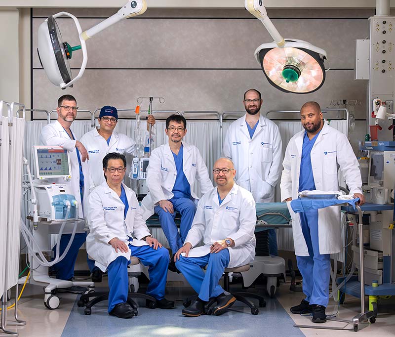 Members of the of the abdominal and heart transplant team are seated and standing in an operating room. Operating room lights are overhead with surgical equipment in the background. 