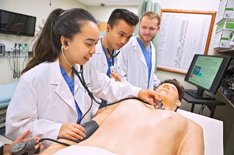 Three medical students wearing lab coats examine a manikin in the Penn State College of Medicine Clinical Simulation Center. A young woman in the front puts a stethoscope on his chest. The man next to her is wearing a stethoscope around his neck, and the man next to him is looking at them. The manikin is lying on an examining table. A monitor is next to them.