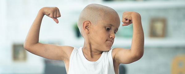 A little boy fighting cancer sits on the sofa at home, with his arms raised and his muscles flexed as he shows how brave he is. He is dressed casually in a white tank top and has his head shaved as he looks down at his muscles.