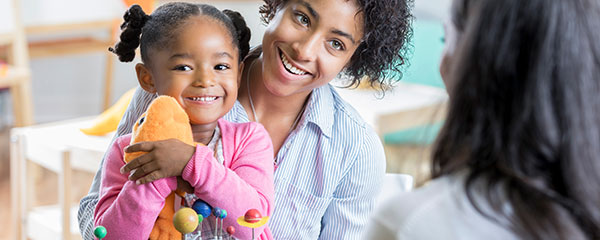 An adorable little preschool age girl sits in her mother's lap in a classroom across the table from her new unrecognizable preschool teacher. She hugs a stuffed animal and plays with a toy as her mother talks to the teacher.