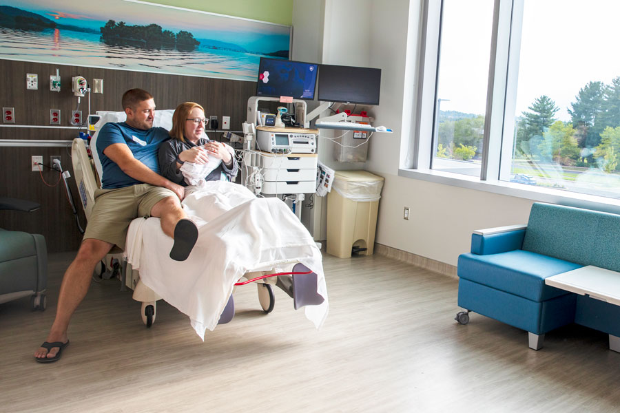 A smiling new mom holds her new baby in a hospital labor and delivery room. The dad is seated on the bed beside her smiling and looking at the baby. The room is spacious and new and has a large set of windows facing trees and mountains.