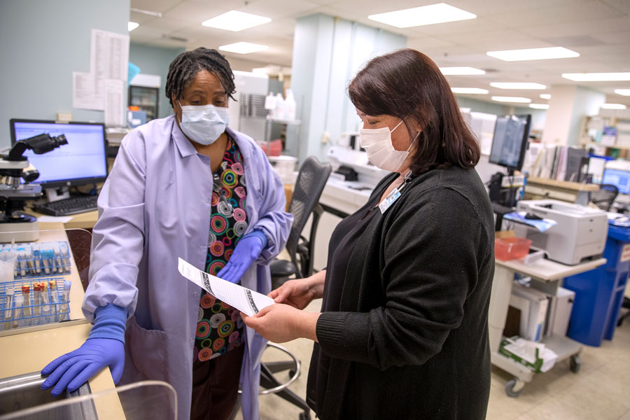 Michelle Murdoch, right, a laboratory manager at Penn State Health Holy Spirit Medical Center, looks at a paper she is holding. She has medium-length hair and is wearing a long-sleeved top and face mask. A woman stands on the left wearing a medical smock, flowered blouse, surgical gloves and a face mask. Test tubes and a microscope are on a counter to their left.