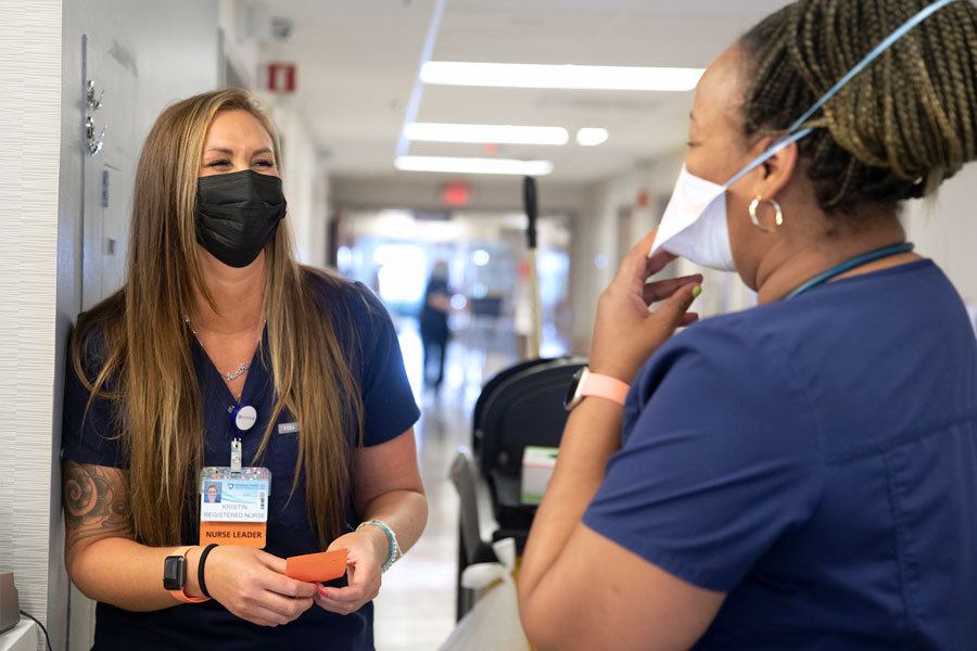 Kristen Hays, left, talks to her team member, Danielle Evans. They are both wearing scrubs and face masks. Hays has long hair and a name badge hanging from her blouse.