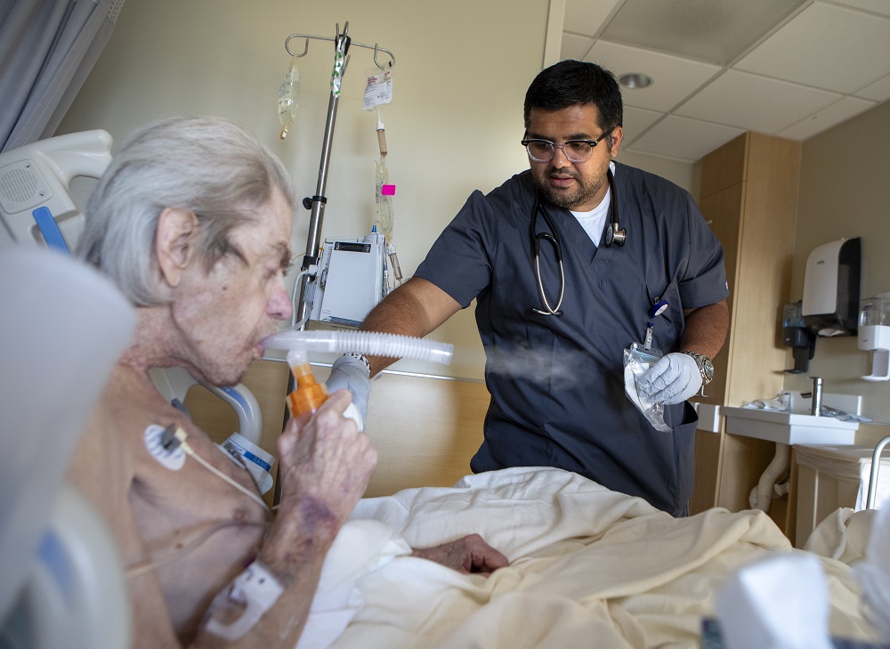 Imran Dawood stands to the right of an older adult male patient, who is sitting up in bed. The patient is breathing into a nebulizer tube. Imran is wearing glasses, is dressed in hospital scrubs and has a stethoscope around his neck. His right arm is extended toward the patient. He holds a small medical bag in his gloved left hand. 