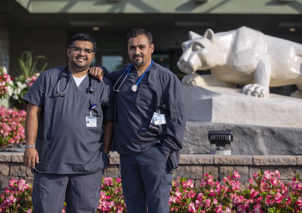 Imran Dawood stands to the left of his brother, Yasin Dawood, outside Penn State Health Holy Spirit Medical Center. They are both smiling, dressed in hospital scrubs and have stethoscopes around their necks and name badges clipped to their pockets. Behind them is a stone wall with flowers at the base of the wall and some to the left of Imran. To their right is a statue of the Penn State Nittany Lion. 