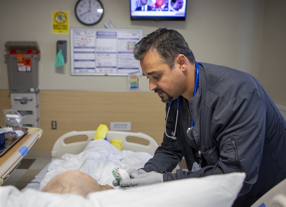 Yasin Dawood, who has a stethoscope around his neck and is wearing a hospital scrub, stands to the right of a patient who is lying in bed. Yasin is bending the patient over to check oxygen levels on a small monitor. A hospital tray is to the left of the patient and a TV screen, clock and hospital staffing chart are on the wall behind the foot of the bed. 