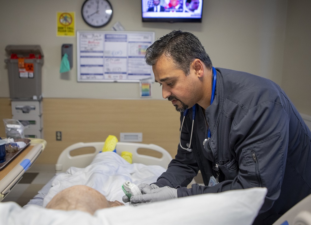 Yasin Dawood, who has a stethoscope around his neck and is wearing a hospital scrub, stands to the right of a patient who is lying in bed. Yasin is bending the patient over to check oxygen levels on a small monitor. A hospital tray is to the left of the patient and a TV screen, clock and hospital staffing chart are on the wall behind the foot of the bed.
