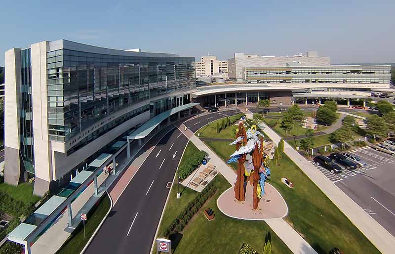 An aerial view of Penn State Children's Hospital (left), Hershey Medical Center (center) and Penn State Cancer Institute (right). A large sculpture is in a grassy area across from the Children’s Hospital. A road and covered walkway are in front of the three buildings.