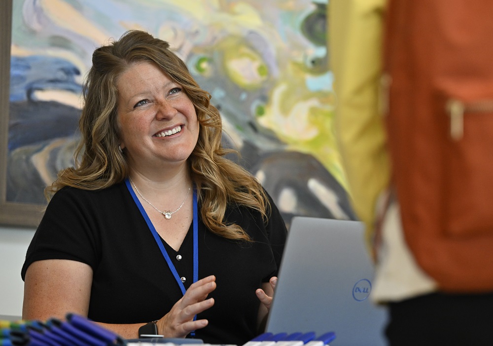 Katy Rush sits at a table at a job fair, smiling at a person standing in front of her and gesturing with her hands. She has an open laptop computer on the table in front of her and a lanyard around her neck. There are giveaways on the table to her right. A painting hangs on the wall behind her.