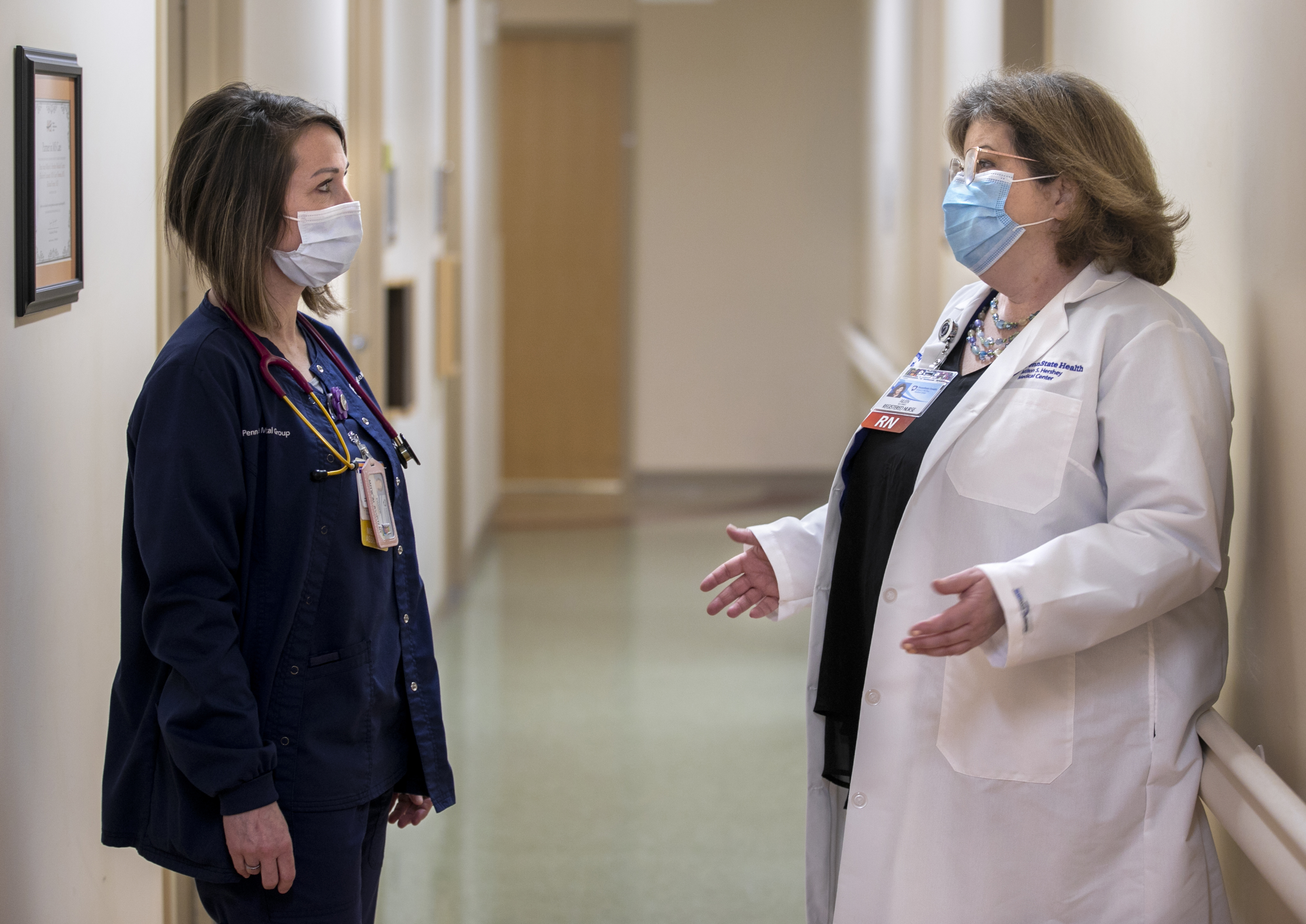 Aileen McCormick, right, gestures as she talks to medical assistant Kimber Sager in a hallway of Hershey Medical Center’s Department of Neurology. McCormick is wearing glasses, a lab coat, name badge and face mask. Sager is wearing a jacket and pants, a face mask and name badge and has a stethoscope around her neck.