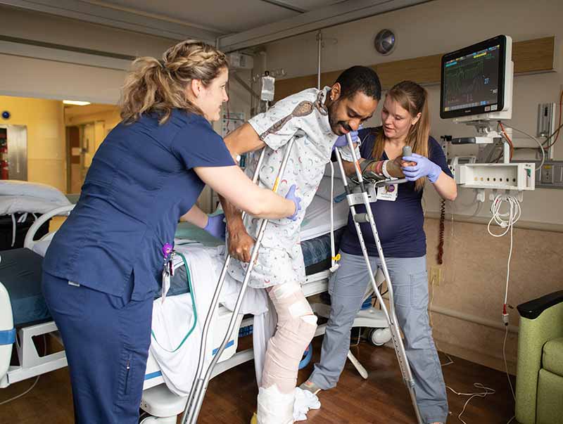 Two nurses hold up a man as he stands using crutches in a hospital room. The nurse on the left has her hair in a braid and is wearing scrubs. The nurse on the right has long hair and is wearing scrubs. The man has his leg bandaged and is wearing a patient smock and has crutches under his arms. A hospital bed is behind them, and a monitor and chair are on the right.
