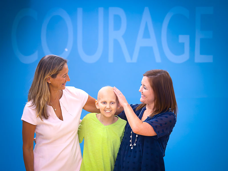 Young child with 2 females standing in front of a blue screen with the word courage being displayed.