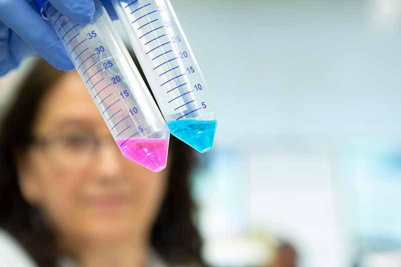 A woman researcher holds two test tubes filled with colorful liquid in front of her face. She has shoulder-length hair and is wearing glasses and surgical gloves. The test tubes have measurement markings.