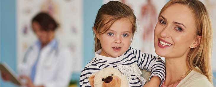 A mother holds her daughter in a hospital room. The girl is holding a stuffed animal.