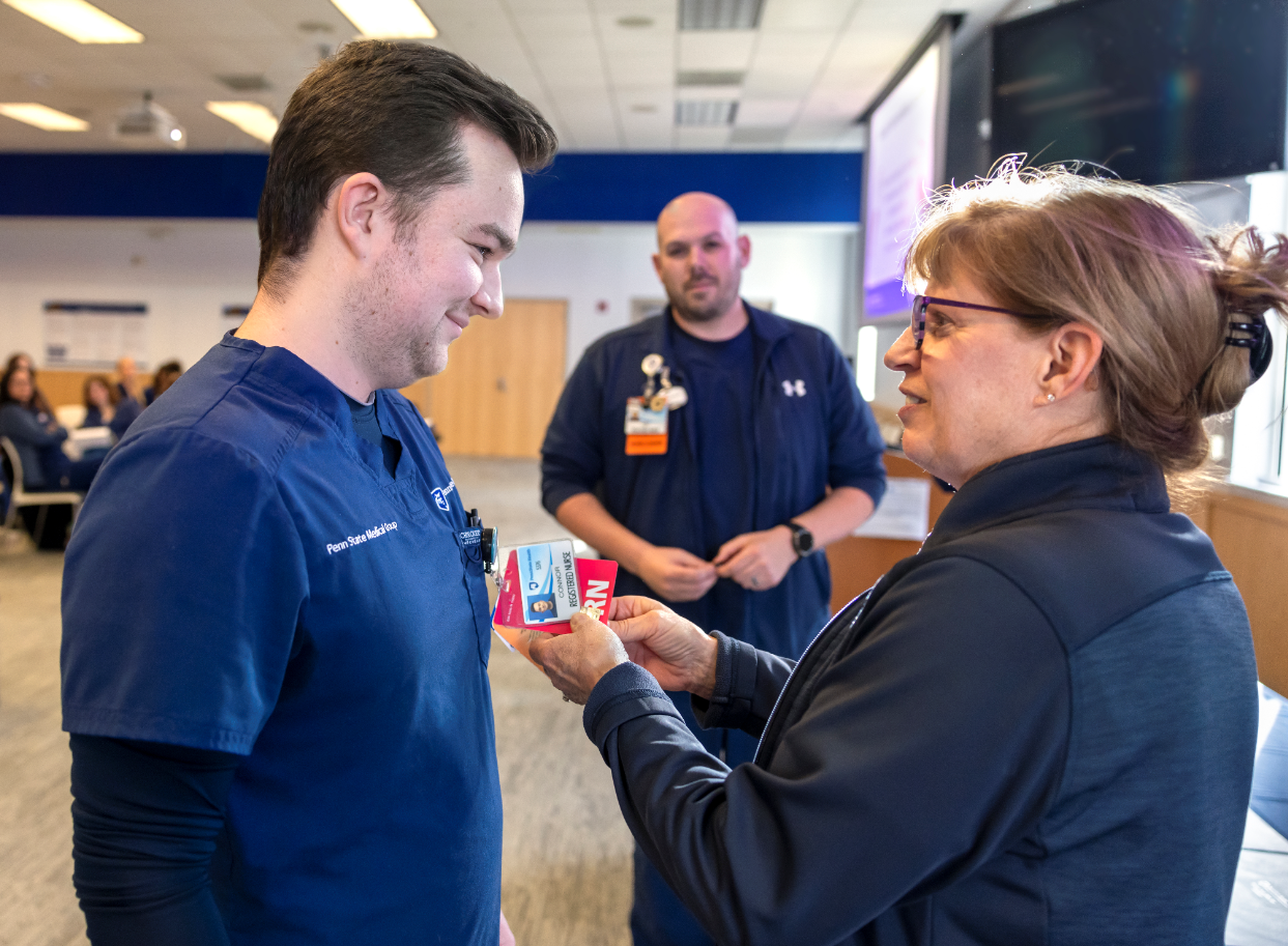 A female registered nurse pins a graduation pin onto a male nurse following the completion of his graduate nurse residency. Both are wearing navy blue uniforms. A male nurse looks on from behind them.