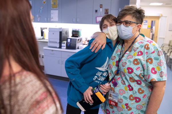 a 15 year old girl, is embraced in a side hug by a registered nurse. Aaliyah has short hair, is wearing a blue sweatshirt and has a mask on her face. The nurse is wearing colorful scrubs and has a mask and glasses on her face. On the left side of the photo, Aaliyah’s mom looks on with only the back of her head and long auburn hair visible
