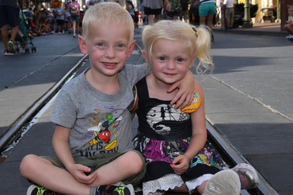 Two small children sit on the ground at Walt Disney World. The boy on the left has his arm around the girl on the right. People can be seen walking and pushing strollers in the background.