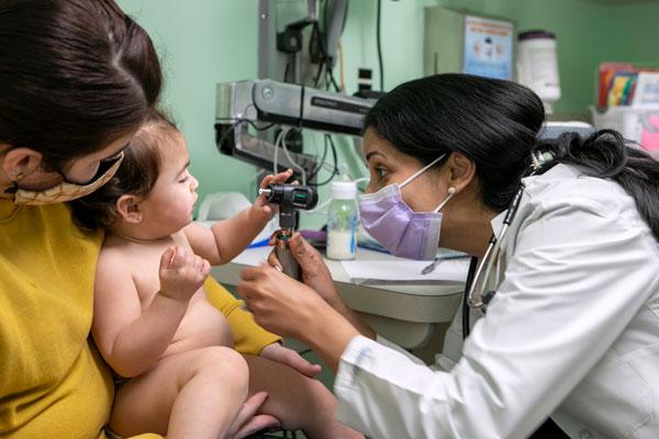 Young mother and infant child sitting in doctors office while physician examining infant with stethoscope.