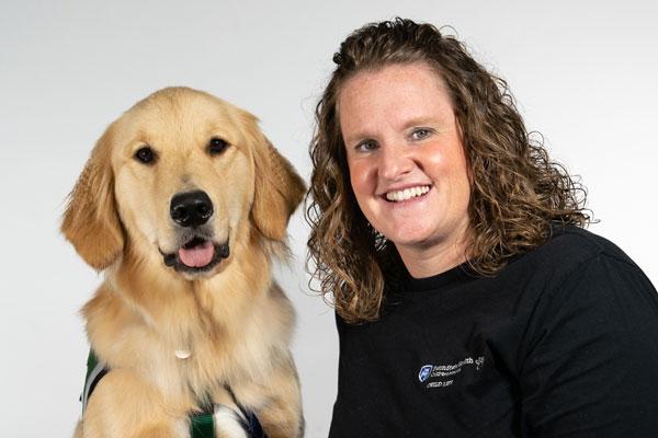A golden retriever dog named Captain is on the left and a women wearing a black top is on the left. The background of the photo is white. Both are looking at the camera. This photo was taken in November 2022.